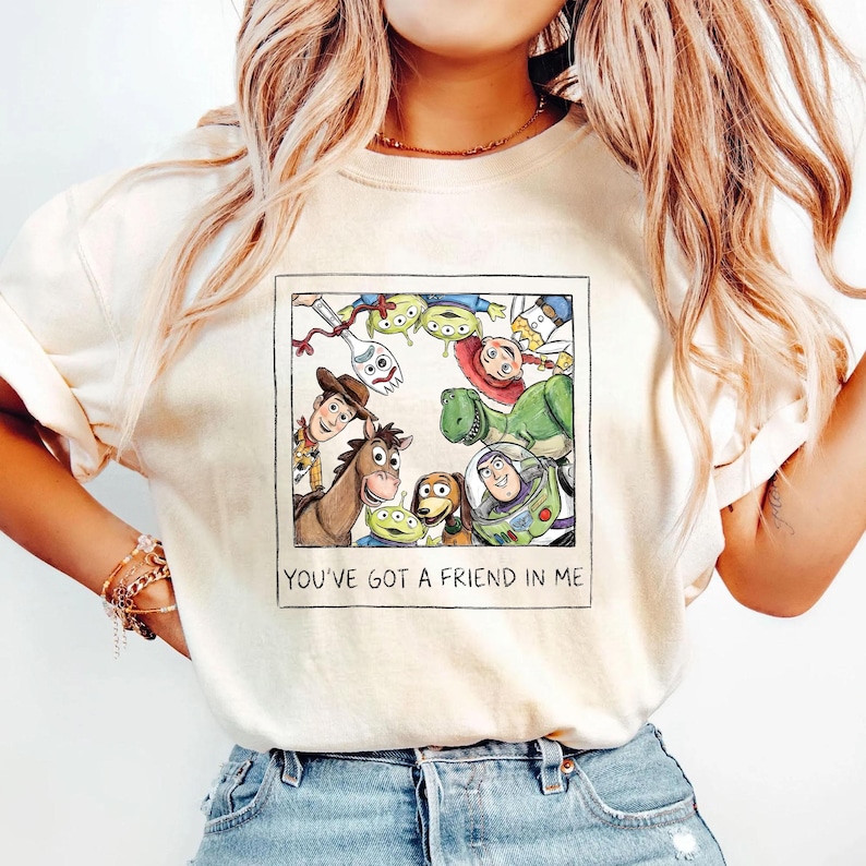 May include: Cream-colored t-shirt featuring a framed illustration of characters from the animated film "Toy Story." The image includes Woody, Buzz Lightyear, and other characters. The text "YOU'VE GOT A FRIEND IN ME" is printed below the image.