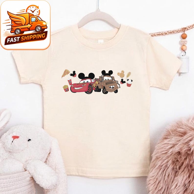May include: A cream-coloured children's t-shirt featuring a graphic of two cartoon cars with Mickey Mouse ears. The red car is on the left, and the brown car is on the right. The shirt also has images of food items.
