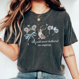 May include: Dark grey t-shirt with a graphic of reindeer and mouse ears with snowflakes and a blue bow. The text "The cold never bothered me anyway" is printed below the graphic. The shirt has a relaxed fit.