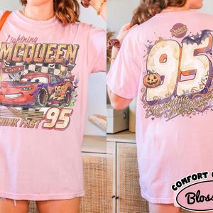 May include: A light pink t-shirt featuring a graphic of Lightning McQueen from Cars on the front with the text Lightning McQueen, Think Fast, and 95. The back displays a large 95 with a pumpkin, a bat, and the text Lightning McQueen.