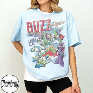 May include: Light blue t-shirt featuring a graphic of Buzz Lightyear from Toy Story. The design includes the words "BUZZ lightyear" and "LASER BLAST" in red and white. The shirt has a vintage, cartoon-like aesthetic.
