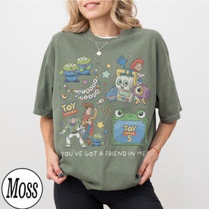 Puede incluir: Una camiseta de manga corta color verde musgo con un estampado gr&aacute;fico de personajes de Toy Story, incluyendo a Woody, Buzz Lightyear y alien&iacute;genas. El dise&ntilde;o incluye el texto Disney Pixar, Toy Story, Toy Story 5 y You've Got a Friend in Me.