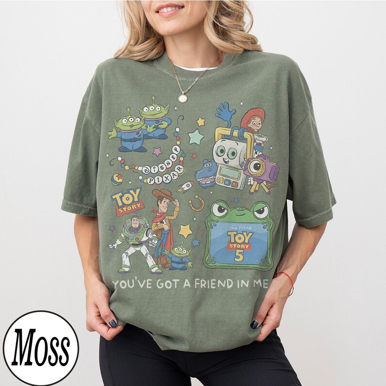 Puede incluir: Una camiseta de manga corta color verde musgo con un estampado gr&aacute;fico de personajes de Toy Story, incluyendo a Woody, Buzz Lightyear y alien&iacute;genas. El dise&ntilde;o incluye el texto Disney Pixar, Toy Story, Toy Story 5 y You've Got a Friend in Me.
