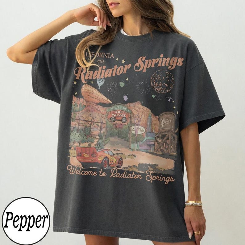 Pu&ograve; includere: T-shirt grigio scuro con una grafica di Radiator Springs, con il testo "California 2012 Radiator Springs" e "Welcome to Radiator Springs". Il design include auto, un paesaggio desertico e fuochi d'artificio.