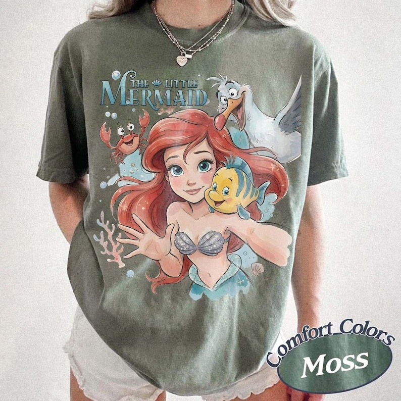 May include: A moss green short-sleeve t-shirt featuring a graphic print of Ariel, Flounder, Sebastian, and Scuttle from The Little Mermaid. The design includes the text The Little Mermaid in blue lettering above the characters with bubbles and coral.
