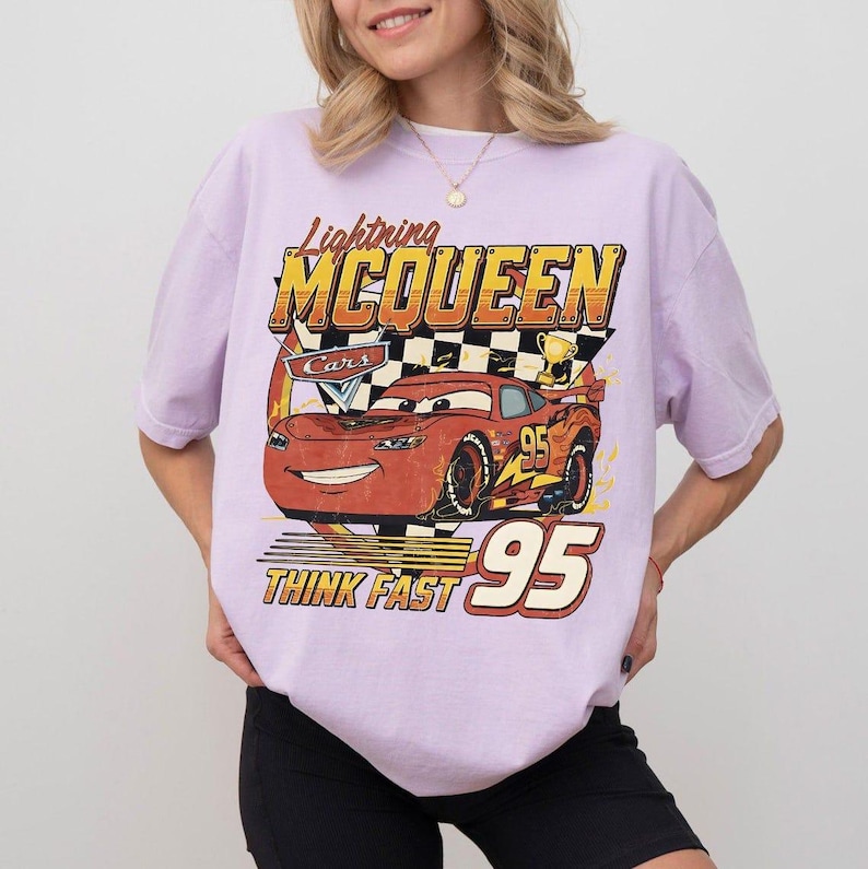 May include: A light purple t-shirt featuring a graphic of Lightning McQueen from the Cars movie. The design includes the text "Lightning McQueen", "Cars", "Think Fast", and the number "95". The car is red with black wheels.