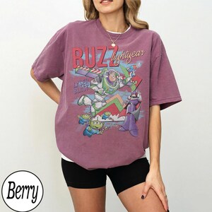 May include: Berry-colored t-shirt featuring a graphic of Buzz Lightyear, aliens, and Zurg from Toy Story. The design includes the text "BUZZ lightyear" and "LASER BLAST". The shirt is a relaxed fit.