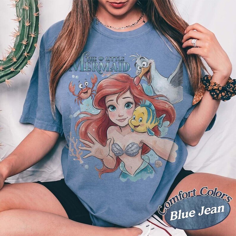 May include: A blue short-sleeve t-shirt featuring a graphic print of Ariel from The Little Mermaid, along with Flounder, Sebastian, and Scuttle. The text The Little Mermaid is printed above the characters in a stylized blue font on the front.
