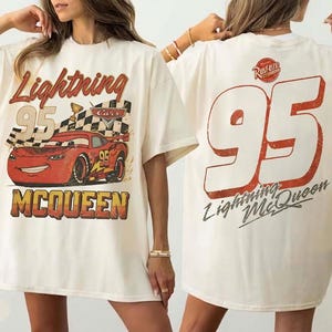 May include: Ivory-colored t-shirt featuring a graphic of Lightning McQueen from the Cars movie. The front displays the car, the number 95, and the name "Lightning McQueen." The back has a large number 95 and the name.