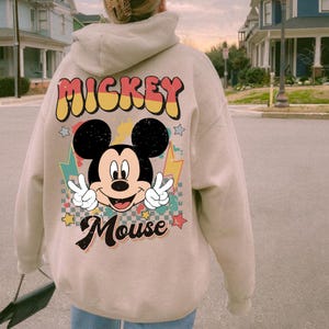 May include: A beige hoodie featuring a graphic of Mickey Mouse with peace signs and the text "Mickey Mouse". The design includes stars and lightning bolts. The hoodie has a drawstring hood and long sleeves.
