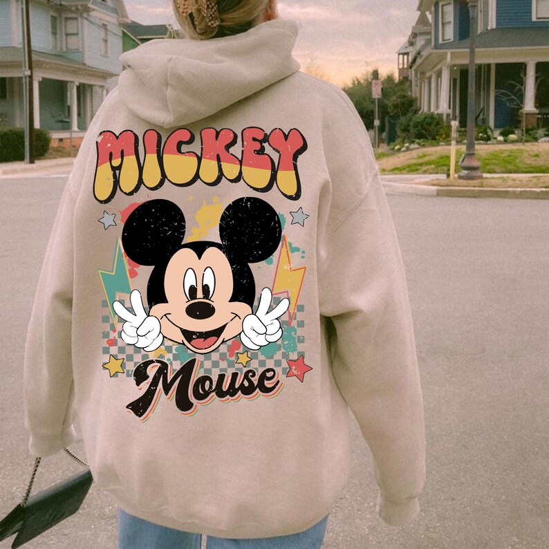 May include: A beige hoodie featuring a graphic of Mickey Mouse with peace signs and the text "Mickey Mouse". The design includes stars and lightning bolts. The hoodie has a drawstring hood and long sleeves.