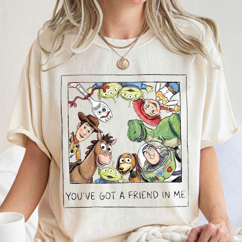 May include: Cream-colored t-shirt featuring a framed illustration of Toy Story characters. The artwork includes Woody, Buzz Lightyear, Rex, and the Little Green Men. The text "YOU'VE GOT A FRIEND IN ME" is printed below the image.
