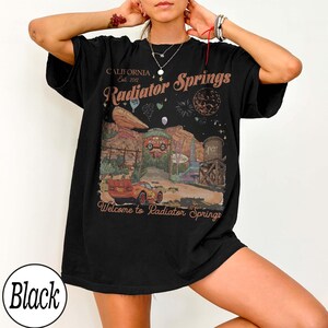 Pu&ograve; includere: T-shirt nera con grafica di Radiator Springs, con testo che recita "California Est. 2012" e "Welcome to Radiator Springs." Il design include un'auto rossa, un paesaggio desertico e una torre dell'acqua.