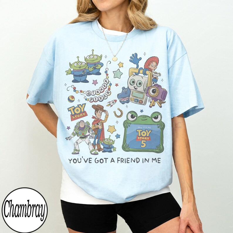 Puede incluir: Una camiseta azul claro con un estampado gr&aacute;fico colorido de personajes de Toy Story, incluyendo a Buzz Lightyear, Woody y alien&iacute;genas. El dise&ntilde;o incluye el texto Disney Pixar, Toy Story 5 y la frase You've Got A Friend In Me.