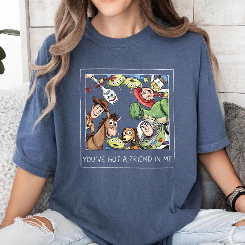 May include: A blue t-shirt featuring a framed illustration of characters from the animated film "Toy Story." The illustration includes Woody, Buzz Lightyear, and other characters. The text "YOU'VE GOT A FRIEND IN ME" is printed below the illustration.