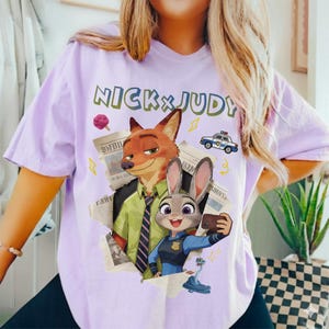 May include: A light purple t-shirt featuring the text "NICK & JUDY" in green letters. The shirt has a graphic of Nick Wilde and Judy Hopps from Zootopia, along with a police car and newspaper clippings.