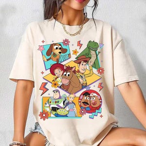 Puede incluir: Camiseta beige con un gr&aacute;fico colorido de personajes de la pel&iacute;cula animada Toy Story. El dise&ntilde;o incluye a Woody, Buzz Lightyear, Rex y otros personajes, con estrellas y flores.