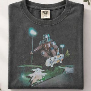May include: Dark gray Comfort Colors t-shirt featuring a graphic of The Mandalorian skateboarding. The image includes Grogu on a skateboard. The shirt has a crew neck and a tag that reads "Comfort Colors".
