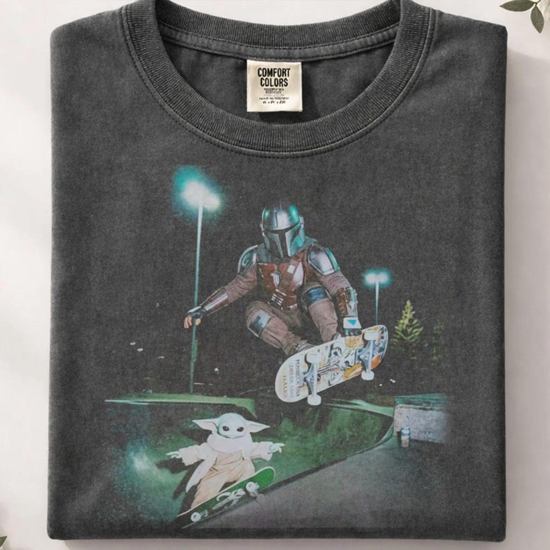 May include: Dark gray Comfort Colors t-shirt featuring a graphic of The Mandalorian skateboarding. The image includes Grogu on a skateboard. The shirt has a crew neck and a tag that reads "Comfort Colors".