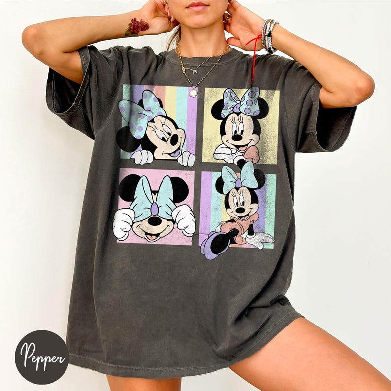 May include: Dark grey t-shirt featuring a four-panel design of Minnie Mouse in various poses. Each panel has a pastel background with vertical stripes. Minnie Mouse is wearing a polka dot bow in each panel. The shirt has a relaxed fit.