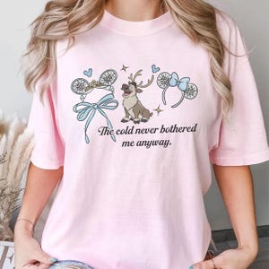 May include: A pale pink t-shirt with a graphic design. The design features a reindeer, mouse ears, snowflakes, and the text "The cold never bothered me anyway." The shirt is made of a soft material.