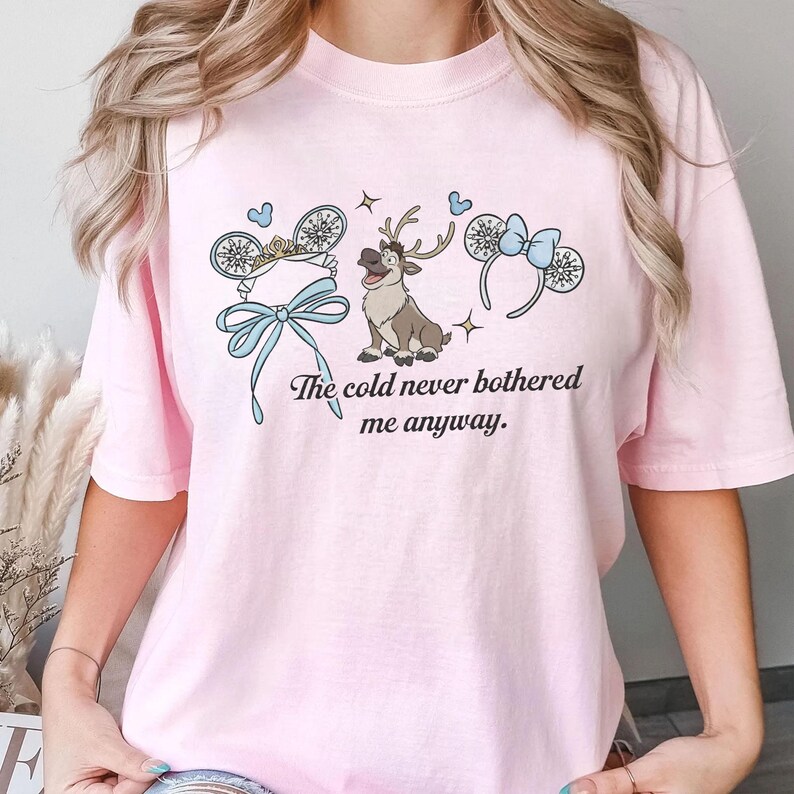 May include: A pale pink t-shirt with a graphic design. The design features a reindeer, mouse ears, snowflakes, and the text "The cold never bothered me anyway." The shirt is made of a soft material.