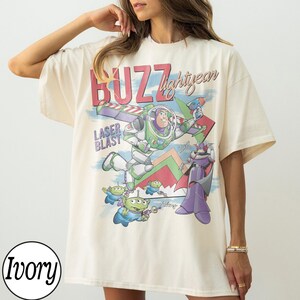 May include: Ivory-colored t-shirt featuring a colorful graphic of Buzz Lightyear, aliens, and Zurg from the movie Toy Story. The shirt has the words "Buzz Lightyear" and "Laser Blast" printed on it. The shirt is a relaxed fit.