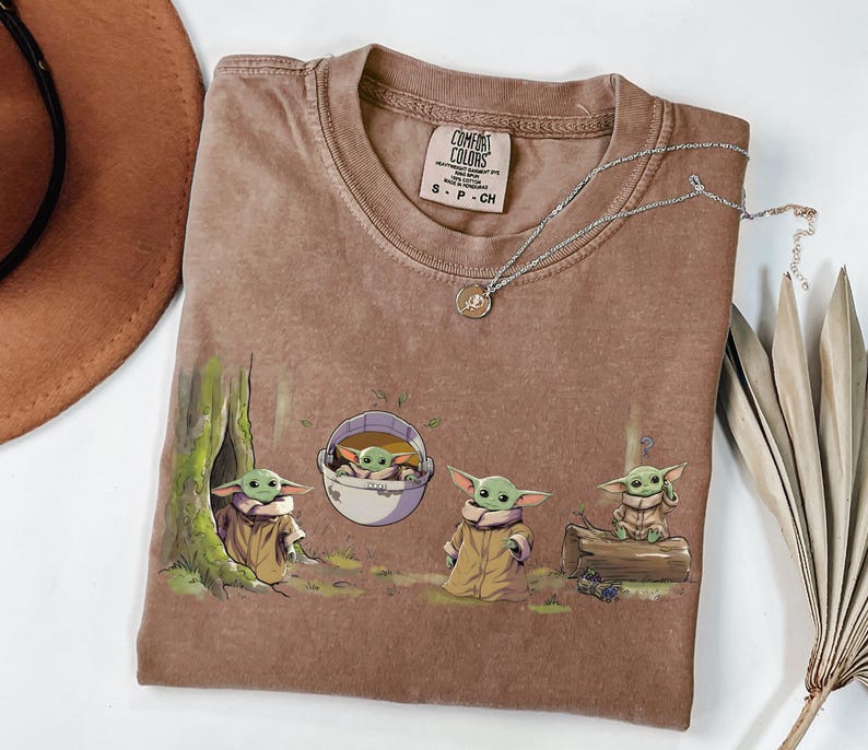May include: A brown short-sleeved t-shirt featuring a graphic print of four small green creatures in various poses, including one inside a floating pod. The shirt has a Comfort Colors brand label at the neck, size small, made in Honduras.
