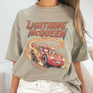 Może przedstawiać: Jasnobrązowy t-shirt z grafiką Zygzaka McQueena, czerwonego samochodu wyścigowego z animowanego filmu Auta. Tekst "LIGHTNING McQUEEN" znajduje się nad samochodem, z błyskawicami i płomieniami w tle.