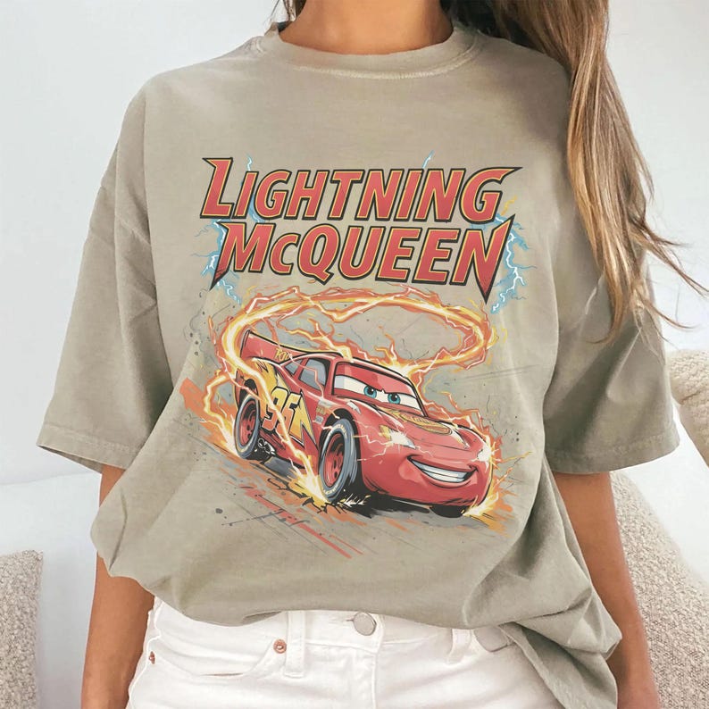 Może przedstawiać: Jasnobrązowy t-shirt z grafiką Zygzaka McQueena, czerwonego samochodu wyścigowego z animowanego filmu Auta. Tekst "LIGHTNING McQUEEN" znajduje się nad samochodem, z błyskawicami i płomieniami w tle.