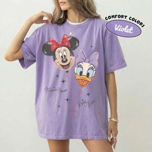 May include: A violet t-shirt featuring a graphic of Minnie Mouse and Daisy Duck as balloons. The Minnie balloon has a red bow, and the Daisy balloon has a purple bow. The text "Minnie Mouse" and "Daisy Duck" is printed below each character.