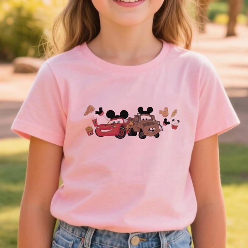 May include: A pale pink t-shirt with a cartoon graphic of two cars with Mickey Mouse ears. A red car and a brown truck are featured. The design includes cartoon food items like pizza and fries.