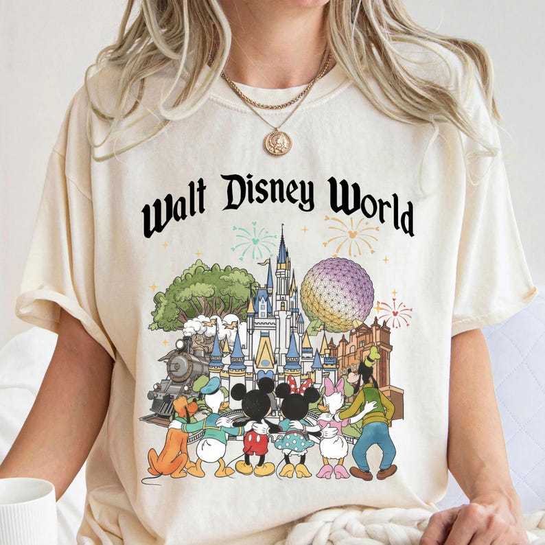 May include: Cream-colored t-shirt featuring a colorful graphic of Disney characters in front of Cinderella's castle, a train, and Spaceship Earth. The words "Walt Disney World" are printed in black script above the graphic. The shirt has short sleeves and a crew neck.
