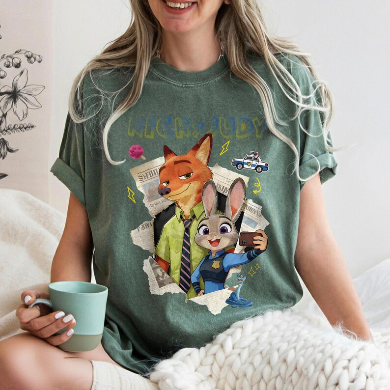 May include: Green t-shirt featuring a graphic of Nick Wilde and Judy Hopps from Zootopia. The design includes the text "Nick & Judy", a police car, and a selfie. The shirt is a relaxed fit.