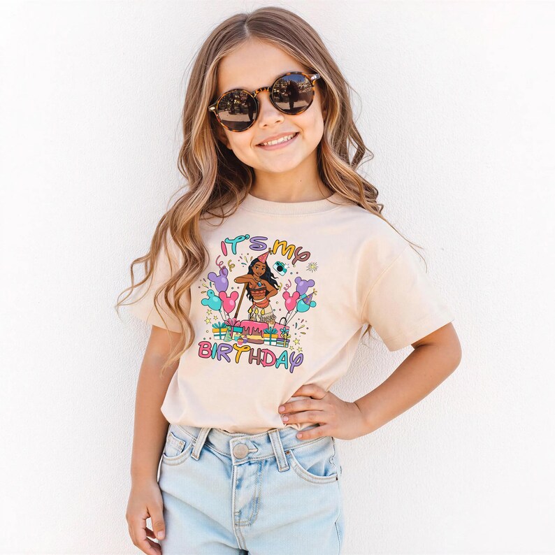 K&ouml;nnte beinhalten: Ein beiges T-Shirt mit einer bunten Grafik mit der Aufschrift "It's my Birthday". Die Grafik zeigt eine Zeichentrickfigur, Luftballons und eine Geburtstagstorte. Die Person tr&auml;gt eine Sonnenbrille und eine hellblaue Jeans.