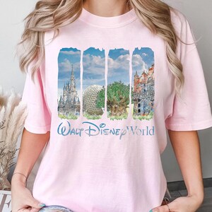 May include: A light pink short-sleeve t-shirt featuring a graphic print of four vertical panels depicting iconic theme park landmarks, including a castle, a geodesic dome, a large tree, and a tower, with the text Walt Disney World below.