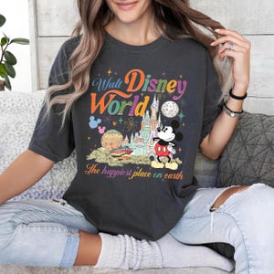 以下が含まれることがあります： ウォルト・ディズニー・ワールドのカラフルなグラフィックがプリントされた濃いグレーのTシャツ。ミッキーマウス、お城、そして「The happiest place on earth」の文字が特徴です。デザインには花火、惑星、星が含まれています。
