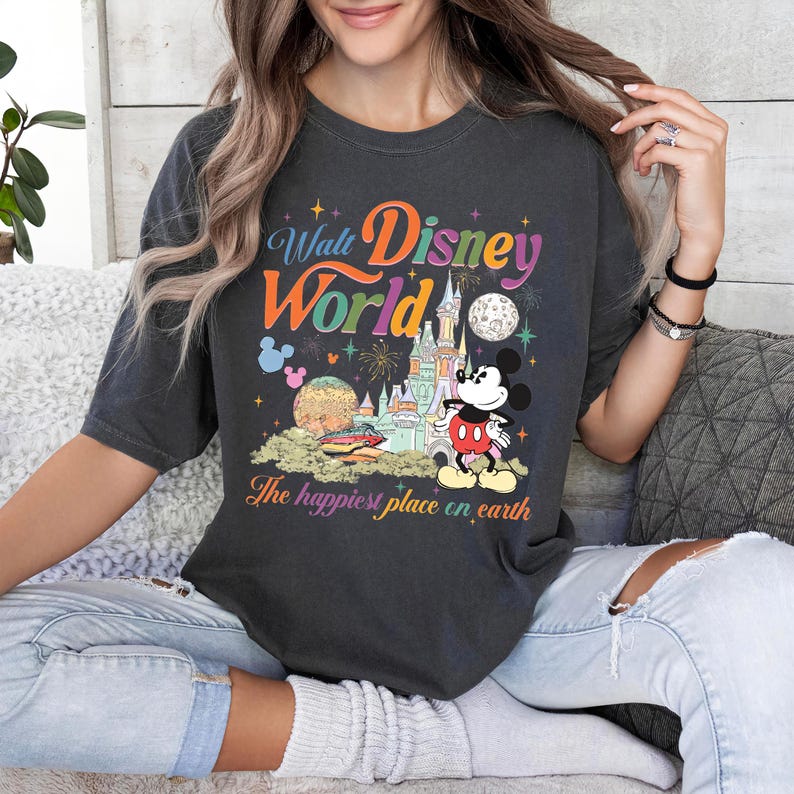 以下が含まれることがあります： ウォルト・ディズニー・ワールドのカラフルなグラフィックがプリントされた濃いグレーのTシャツ。ミッキーマウス、お城、そして「The happiest place on earth」の文字が特徴です。デザインには花火、惑星、星が含まれています。