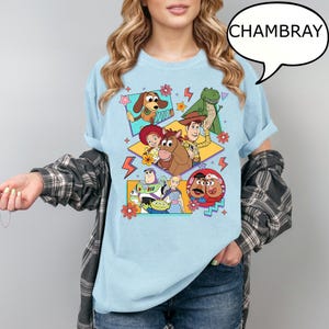 Puede incluir: Camiseta azul claro con un dise&ntilde;o gr&aacute;fico colorido de personajes de Toy Story. El dise&ntilde;o incluye a Woody, Buzz Lightyear y otros personajes. La palabra "CHAMBRAY" est&aacute; en una burbuja.