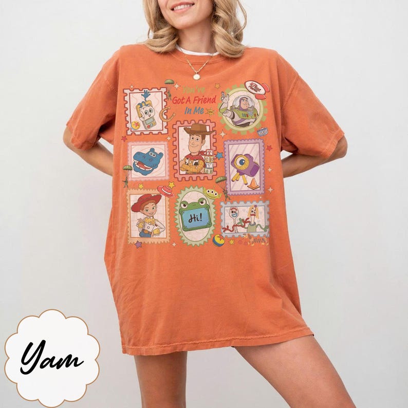 May include: An orange short-sleeve t-shirt featuring a graphic print of various cartoon characters in postage stamp-style frames. The central text reads "You've Got A Friend In Me" surrounded by illustrations of a cowboy, space ranger, and other toys.