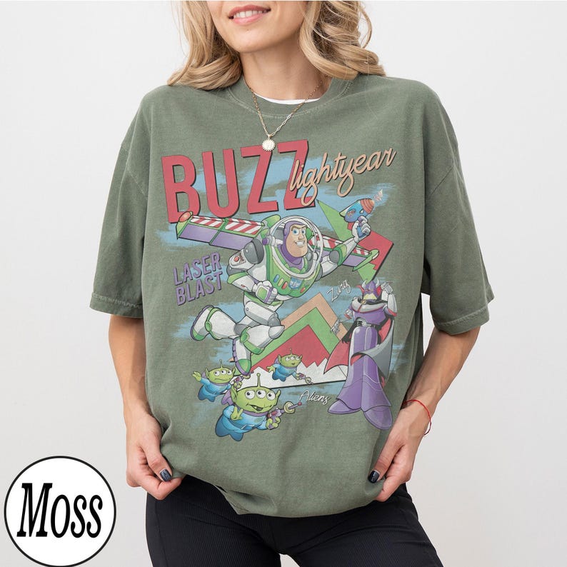 May include: Olive green t-shirt featuring a graphic of Buzz Lightyear, aliens, and Zurg from Toy Story. The design includes the text "BUZZ lightyear" and "LASER BLAST".