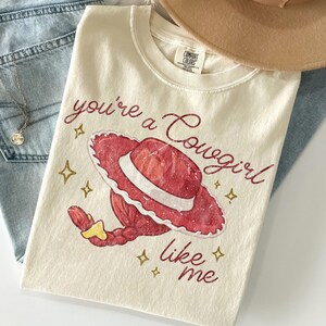 May include: A cream-colored short-sleeve t-shirt featuring a graphic of a red cowgirl hat and a red braided ponytail. The text reads, "you're a Cowgirl like me," accompanied by small gold star icons. The shirt has a Comfort Colors brand label.