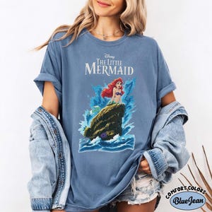 May include: A blue short-sleeve t-shirt featuring a graphic of Ariel from Disney's The Little Mermaid sitting on a rock amidst blue ocean waves. The text Disney The Little Mermaid is printed in white above the character. The shirt has a crew neckline.