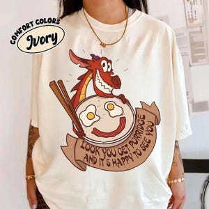 Op de afbeelding: Ivoor T-shirt met een cartoon draak die een bord eieren en spek met eetstokjes vasthoudt. Het bord heeft een lachend gezicht. De tekst op het shirt luidt "LOOK YOU GET PORRIDGE AND IT'S HAPPY TO SEE YOU."