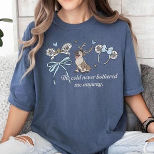 May include: A blue t-shirt with a cartoon reindeer, snowflake-themed mouse ears, and the text "The cold never bothered me anyway." The design incorporates light blue bows and accents, giving a whimsical and playful appearance.