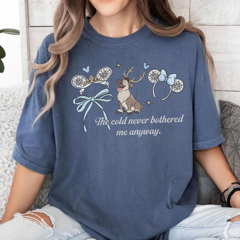 May include: A blue t-shirt with a cartoon reindeer, snowflake-themed mouse ears, and the text "The cold never bothered me anyway." The design incorporates light blue bows and accents, giving a whimsical and playful appearance.
