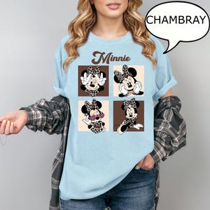 May include: Light blue t-shirt featuring a Minnie Mouse graphic. The graphic includes four panels with different poses of Minnie Mouse wearing a leopard print bow and dress. The word "Minnie" is printed above the panels in a brown script font.
