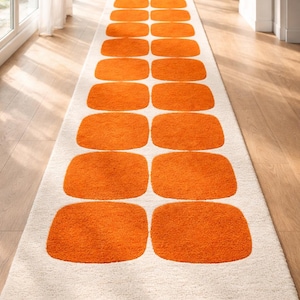 Könnte beinhalten: Ein langer, rechteckiger Teppich mit cremefarbenem Hintergrund und einem sich wiederholenden Muster aus großen, orangefarbenen, abgerundeten Rechtecken. Der Teppich liegt auf einem Holzboden und ist ein modernes Designelement.