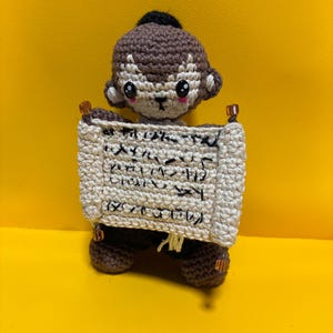 May include: Crocheted monkey doll holding a scroll with Japanese text. The monkey is brown with a black topknot and white face details. The scroll is off-white with black text and brown accents. The doll is on a yellow background.