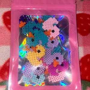 Pode incluir: Uma bolsa rosa, selável, contendo uma coleção de contas coloridas em forma de flor pixelizada. As contas são de várias cores, incluindo azul, roxo, rosa, amarelo e laranja. A bolsa tem um interior holográfico iridescente.